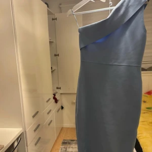 Ljusblå one shoulder klänning - Snygg ljusblå klänning med one shoulder-design och volang nertill. Klänningen har en synlig dragkedja på baksidan och är figurnära med en stilren look. Perfekt för dig som vill sticka ut med en modern och trendig stil.