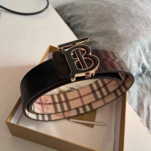Burberry rutigt skärp med guldspänne - Säljer ett snyggt Burberry-bälte i skinn med klassiskt rutigt mönster i beige, brun, svart och röd. Bältet har ett stort guldigt spänne med Burberrys ikoniska TB-logga och är vändbart med både mörk och ljus sida. Perfekt accessoar för att lyfta din outfit.