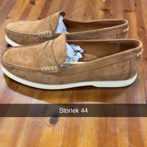 Bruna mockaloafers Polo Ralph Lauren - Snygga bruna loafers från Polo Ralph Lauren i mjuk mocka med klassisk design och vita kontrastsömmar. Skorna har en platt sula och diskret logga på sidan. Perfekta för en stilren och avslappnad look.