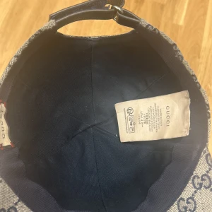 Gucci keps med GG-mönster och röd-blå rand - Snygg keps från Gucci i beige och marinblått GG-monogram. Kepsen har en klassisk böjd skärm, justerbart läderband bak och en ikonisk röd-blå rand på sidan. Tillverkad i bomullscanvas med detaljer i skinn. Perfekt för dig som vill sticka ut med lyxig streetstyle. Pris kan diskuteras