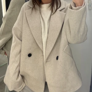 Beige oversized kavajjacka med ull - Snygg beige kavajjacka i oversized modell med breda slag och två svarta knappar framtill. Jackan har fickor på sidorna och är tillverkad i ett mjukt ullmaterial som ger en lyxig känsla. Perfekt för dig som gillar en stilren och trendig look.