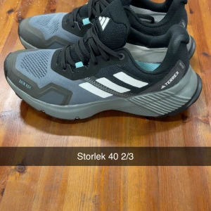 Adidas Terrex sneakers grå/svarta - Säljer ett par Adidas Terrex sneakers i grått och svart med vita ränder. Skorna har snörning, robust sula och meshdetaljer för extra ventilation. Perfekta för dig som gillar en sportig och modern look.