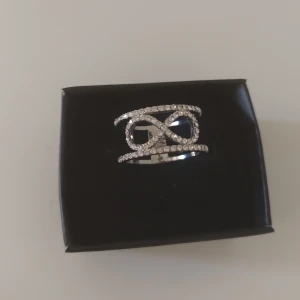Glittrig silverring med stenar - Snygg bred silverring med glittriga stenar i ett coolt oändlighetsmönster. Ringen har en öppen design med två parallella band och gnistrande stenar som täcker hela framsidan. Perfekt för dig som gillar statement-smycken och vill sticka ut.