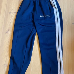 Palm Angels blå trackpants med vita ränder - Snygga blå trackpants från Palm Angels med vita och svarta ränder längs sidorna. Byxorna har elastisk midja med snörning, sidofickor och dragkedja vid benslut. Loggan är tryckt framtill och materialet känns mjukt och sportigt.