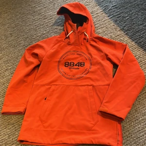 Orange vindjacka från 8848 Altitude - Snygg orange vindjacka från 8848 Altitude med stor logga på bröstet. Jackan har huva med snörning, stor magficka och långa ärmar. Insidan är fodrad med mjuk fleece för extra komfort. Perfekt för dig som gillar outdoor och streetstyle.