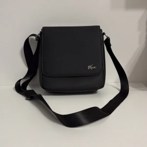 Svart axelväska från Lacoste - Stilren svart axelväska från Lacoste med klassisk krokodillogga i metall på framsidan. Väskan har justerbar axelrem, lock med magnetstängning och ett huvudfack med dragkedja. Tillverkad i slitstark syntet med minimalistisk design och diskreta detaljer.