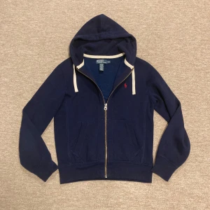Marinblå hoodie från Polo Ralph Lauren - Marinblå hoodie från Polo Ralph Lauren med dragkedja, huva med vita snören och klassisk röd broderad logga på bröstet. Tillverkad i mjuk bomullsblandning med fickor framtill. Perfekt för en chill och stilren look. Passar 170-175 cm