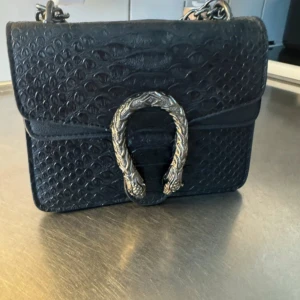 Svart axelväska med kedja och ormdetalj - Snygg svart axelväska med präglat ormmönster och en stor silverfärgad spänne framtill. Väskan har en grov metallkedja som axelrem och flera fack inuti. Perfekt storlek för det viktigaste och ger en cool vibe till din outfit.