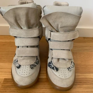 Beige Isabel Marant wedge sneakers - Säljer ett par ikoniska wedge sneakers från Isabel Marant i beige mocka med svarta och vita mönstrade detaljer. Skorna har tre kardborreband, rund tå och gummisula. Perfekt statement-piece med cool design och sköna material.