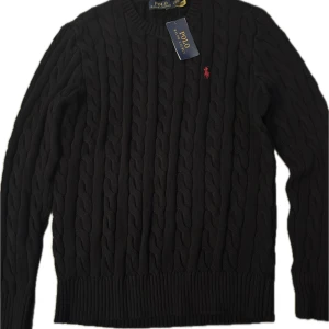 Svart kabelstickad tröja Polo Ralph Lauren - Svart kabelstickad tröja från Polo Ralph Lauren med klassisk röd broderad logga på bröstet. Tröjan har rund hals och ribbade muddar. Perfekt för dig som gillar stilrena och tidlösa plagg med premiumkänsla. Kan släppas smidigare vid snabb affär..😉