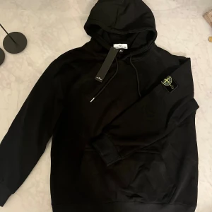 Svart hoodie från Stone Island M - Svart hoodie från i storlek M med klassisk logotyp-patch på ärmen. Tröjan har huva med snörning, ribbade muddar och en stor magficka. Materialet är mjuk bomull och passformen är normal. Perfekt för dig som gillar streetwear och snygga detaljer.