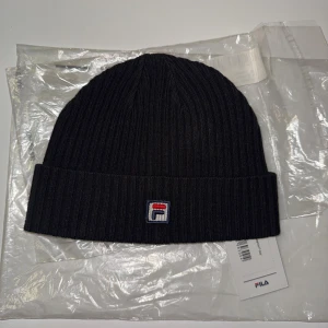Svart ribbad mössa från FILA - Svart ribbstickad mössa från FILA med uppvikt kant och klassisk FILA logga framtill. Snygg och enkel design som passar till allt, tillverkad i mjukt material för extra komfort. Perfekt accessoar för kyliga dagar. Helt ny med prislappen kvar.