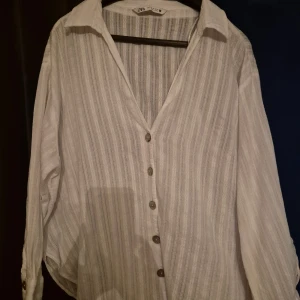 Vit randig skjorta från Zara XL - Vit skjorta från Zara i storlek XL med tunna vertikala ränder och snygga knappar framtill. Skjortan har klassisk krage, lång ärm och är gjord i ett lätt bomullsmaterial som känns luftigt. Perfekt för dig som gillar stilrena och fräscha plagg.