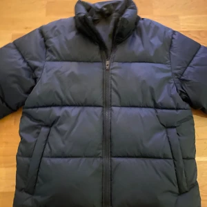 Svart pufferjacka från Everest - Svart pufferjacka i stil från Everest med hög krage och dragkedja framtill. Perfekt för kalla dagar och har en klassisk, loose passform som ger skön komfort. Inbyggd Reflex finns i en av jackfickorna.