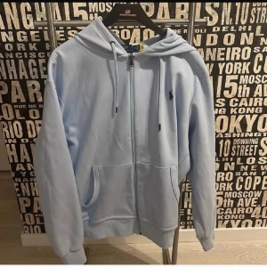 Ljusblå hoodie från Polo Ralph Lauren - Säljer en ljusblå hoodie från Polo Ralph Lauren med dragkedja, huva och snörning. Tröjan har två stora fickor framtill och den klassiska broderade loggan på bröstet. Perfekt för chill dagar och snygg att matcha med jeans.