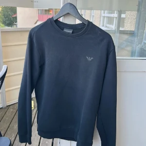 Svart sweatshirt från Emporio Armani - Snygg svart sweatshirt från Emporio Armani med diskret logga på bröstet. Tröjan har rund halsringning och långa ärmar, perfekt för en clean och stilren look. Tillverkad i mjuk bomull som känns skön mot huden. Liten i storken 