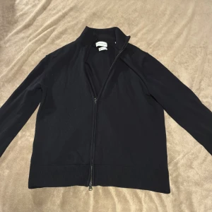 Svart merinoullskofta från These Glory Days - Svart kofta i merinoull från These Glory Days. Klassisk modell med hel dragkedja framtill, ribbade muddar och hög krage. Perfekt för lager-på-lager och riktigt skön känsla tack vare merinoullen. Enkel och stilren design som funkar till allt. Ändast en liten skrapa på axeln, men inget märkvärdigt.