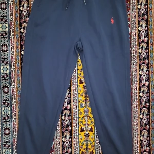 Mörkblå mjukisbyxor Polo Ralph Lauren - Snygga mörkblå mjukisbyxor från Polo Ralph Lauren med klassisk röd logga broderad på vänster höft. Byxorna har snörning i midjan och är gjorda i mjukt bomullsmaterial för extra komfort. Perfekt för chill dagar eller när du vill vara extra bekväm. Den kanske ser stor ut med den är i slim-fit så de passar också för de som har 1-2 storlekar mindre