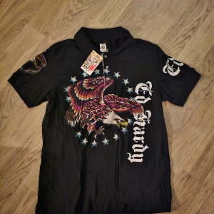 Svart Ed Hardy henley med örntryck - Svart henley från Ed Hardy med stort färgglatt örntryck och stjärnor framtill, samt logga på både fram- och baksida. Skjortan har korta ärmar och detaljer i vitt och flera färger.
