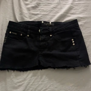 Svart jeanskjol från Marc Lauge - Svart kort jeanskjol från Marc Lauge med rå kant nedtill och coola nitar på fickorna. Baksidan har broderade detaljer på fickorna och kjolen har klassisk jeanslook med knapp och dragkedja framtill.