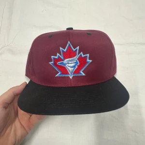 Vinröd snapback keps med broderi - Snygg vinröd snapback keps med svart skärm och grönt under visiret. Framtill finns ett broderat motiv med ett rött lönnlöv och blå detaljer. Justerbar baktill med klassisk snapback-spänne. Perfekt för dig som vill sticka ut med en unik keps.