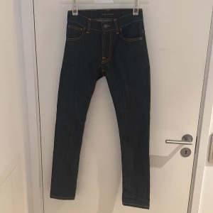Mörkblå jeans från Nudie Jeans, 26/30 - Snygga mörkblå jeans från Nudie Jeans med klassisk orange kontrastsöm och ikonisk broderad detalj på bakfickorna. Modellen har smal passform och raka ben, perfekt för dig som gillar stilrena och tidlösa jeans. Materialet är slitstarkt denim.