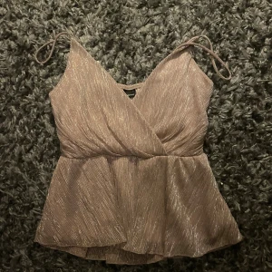 Glittrig omlottopp från Gina Tricot - Supersnygg glittrig topp från Gina Tricot i ljus champagnefärg. Omlottmodell med smala axelband och lätt utsvängd nederdel. Perfekt för dig som vill sticka ut med en feminin och trendig look. Materialet är glansigt och ger en lyxig känsla.