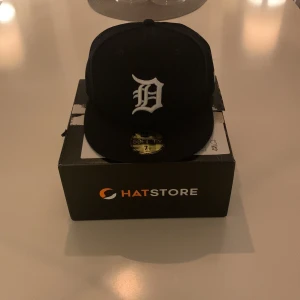 Svart New Era 59FIFTY Detroit Tigers keps - Säljer en mörkblå New Era 59FIFTY fitted keps med Detroit Tigers-logga broderad i vitt framtill och MLB-logga bak. Klassisk rak skärm och hög profil. Kepsen är tillverkad i bomull och polyester, perfekt för dig som gillar streetwear och sportig stil. Helt ny direkt från kartongen, aldrig använd pga att den inte passa.