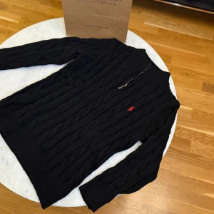 Svart kabelstickad tröja Polo Ralph Lauren - Svart kabelstickad tröja från Polo Ralph Lauren med halv dragkedja och klassisk röd logga broderad på bröstet. Tröjan har ribbade muddar och hög krage, perfekt för kyliga dagar. Materialet är mjuk ull och passformen är normal.