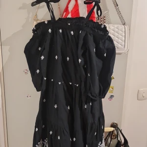 Svart offshoulder klänning med broderi - Svart offshoulder klänning med vita broderade detaljer och knytband på axlarna. Klänningen har puffiga trekvartsärmar och vacker broderad kant längst ner. Tillverkad i mjuk bomull, perfekt för sommarens varma dagar.