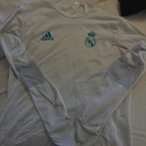 Vit långärmad Real Madrid Adidas tröja - Säljer en vit långärmad tröja från Adidas med Real Madrid-logga och Adidas-tryck i turkos på bröstet. Tröjan har diskreta grå detaljer på armbågarna och är gjord i mjukt polyester-material. Perfekt för fotbollsträning eller chill.