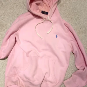Rosa hoodie från Polo Ralph Lauren - Säljer en ljusrosa hoodie från Polo Ralph Lauren med klassisk huva och snörning. Tröjan har en stor ficka framtill och det ikoniska blå Polo-logot broderat på bröstet. Perfekt för chill dagar och enkel att matcha med jeans eller joggers.