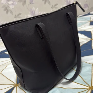 Svart axelväska i skinn från Adfield - Stilren svart axelväska från Anna field i slätt skinnmaterial. Väskan har en klassisk rektangulär form med långa axelremmar och dragkedja upptill. Insidan har ett fack med dragkedja och flera mindre fack för småsaker. Perfekt för dig som gillar minimalistisk design.