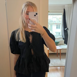Svart blus med puffärm och knyt - Svart blus med korta puffärmar och tre knyt framtill. Blusen har rund hals och volang nedtill som ger en cool och feminin siluett. Materialet känns som bomull och toppen är lätt och luftig, perfekt för att styla med jeans eller kjol.