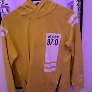 Gul hoodie från Woxo  - Säljer en gul hoodie från Woxo 720 med coolt tryck 'WX CREW 87.0' på bröstet och vita ränder på ärmarna. Tröjan har huva, långa ärmar och dragkedjor vid fickorna. Perfekt för dig som gillar streetstyle och vill sticka ut.