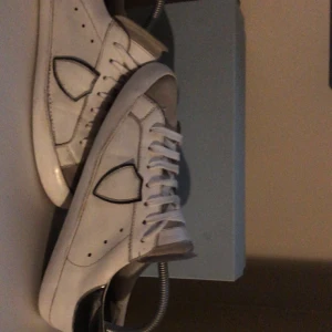 Philippe Model vita sneakers junior - Snygga vita sneakers från Philippe Model Junior med svarta detaljer och svart hälparti. Skorna har klassisk snörning, rund tå och är tillverkade i skinn. Perfekta för dig som vill ha en clean och stilren look med en exklusiv touch. Priset inte huget i sten