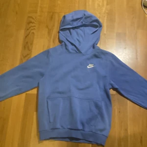 Blå hoodie från Nike med broderad logga - Blå hoodie från Nike med broderad vit logga på bröstet. Tröjan har huva, lång ärm och en stor magficka framtill. Tillverkad i mjuk bomullsmix som är skön att ha på sig. Perfekt för chill eller träning.
