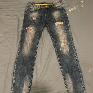 Bruno Leoni slitna jeans med detaljer - Säljer ett par coola mörkblå jeans från Bruno Leoni med slitna och trasiga detaljer samt inslag av gul och röd färg i hålen. Jeansen har klassisk femficksdesign, gul knapp och gul insida i midjan. Materialet är bomull och polyester.