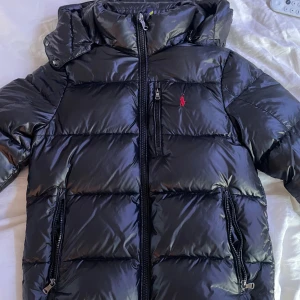 Svart pufferjacka från Polo Ralph Lauren - Snygg svart pufferjacka från Polo Ralph Lauren med glansig finish och röd logga på bröstet. Jackan har huva, dragkedja framtill och två dragkedjefickor. Perfekt för kalla dagar och ger en clean, modern look. Defektfri, kvitto från NK finns!