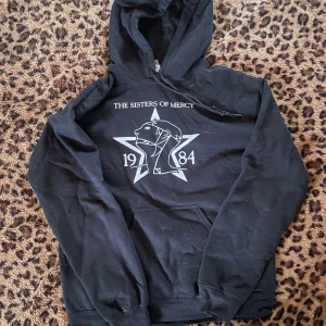 Svart Sisters of Mercy hoodie - Svart hoodie med tryck från The Sisters of Mercy, både fram och bak. Stor vit stjärna och grafiskt ansikte med texten 1984 på framsidan. Klassisk huva och känguruficka. Perfekt för dig som gillar bandmerch och vill sticka ut.