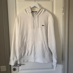 Vit hoodie från Lacoste med dragkedja - Vit hoodie från Lacoste i klassisk passform, med broderad krokodil på bröstet. Tröjan har huva med snörning, två fickor fram och hel dragkedja. 