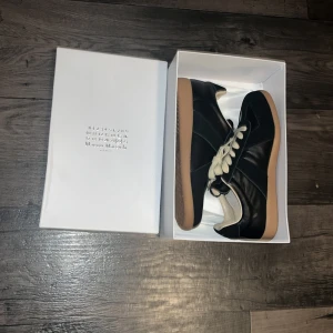 Svarta sneakers från Maison Margiela - Maison Margiela sneakers i svart skinn med beige snörning och gummisula. Klassisk och stilren design med rund tå och platt sula. Perfekta för dig som gillar minimalistisk stil och exklusiva märken.