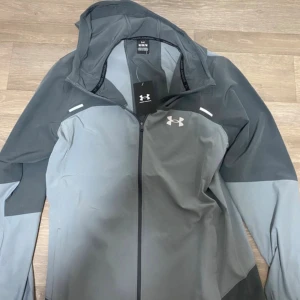 Grå vindjacka från Under Armour - Snygg grå vindjacka från Under Armour med huva och hel dragkedja framtill. Jackan har en cool tvåtonad design med logga på bröstet och stor Under Armour-text på ryggen. Perfekt för träning eller chill ute. Långärmad och tillverkad i lätt polyester.