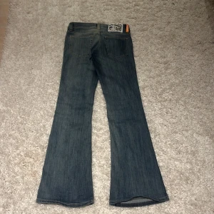 Cheap Monday blå bootcut jeans - Säljer ett par blå bootcut jeans från Cheap Monday med klassisk femficksdesign och ljus tvätt. Jeansen har en snygg logopatch bak och guldiga detaljer vid knapparna. Perfekt för dig som gillar retrovibbar och breda ben.