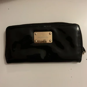 Svart plånbok från Puccini med dragkedja - Stilren svart plånbok från Puccini i glansigt material med guldfärgad metallbricka framtill. Plånboken har dragkedja runtom och insida i röd satin med flera kortfack och myntfack. Perfekt för dig som vill ha en snygg och praktisk accessoar. Lite repig som man ser på bilden med blixten men fungerar utmärkt.