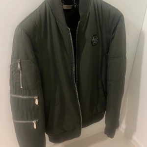 Mörkgrön bomberjacka från Philipp Plein - Snygg mörkgrön bomberjacka från Philipp Plein med silverfärgade dragkedjor på ärmen och framtill. Jackan har ribbade muddar, emblem på bröstet och flera fickor. Perfekt för dig som vill ha en cool och urban stil.