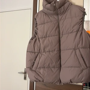 Beige quiltad dunväst från 157 - Snygg beige dunväst från 157 i storlek M. Västen har hög krage, dragkedja framtill och två praktiska fickor med tryckknapp. Quiltad design ger en cool och puffig look, perfekt att slänga över hoodien när det är kyligt ute.