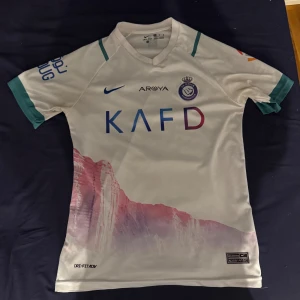 Al Nassr Ronaldo bortatröja Nike S - Säljer en officiell Al Nassr bortatröja med Ronaldo 7 på ryggen. Tröjan är vit med gröna detaljer på ärmarna, tryck i blått och lila samt coolt bergsmotiv nertill. Tillverkad i Dri-FIT-material som andas, perfekt för fotboll eller träning.