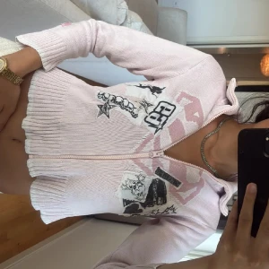 Ljusrosa stickad kofta med tryck - Säljer en ljusrosa stickad kofta med dragkedja och coola svarta och vita tryck framtill. Koftan är croppad med bred ribbad kant och långa ärmar. Perfekt för dig som gillar unika detaljer och streetstyle. Tajt 