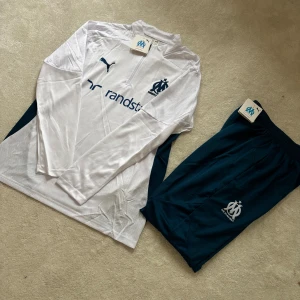Marseille långärmad fotbollströja Puma - Säljer en vit långärmad fotbollströja från Puma med Olympique de Marseille-logga och sponsortryck i marinblått. Tröjan har halv dragkedja fram, detaljer i mörkblått på sidorna och är gjord i 100% polyester med DryCell-teknologi. Perfekt för träning eller match!